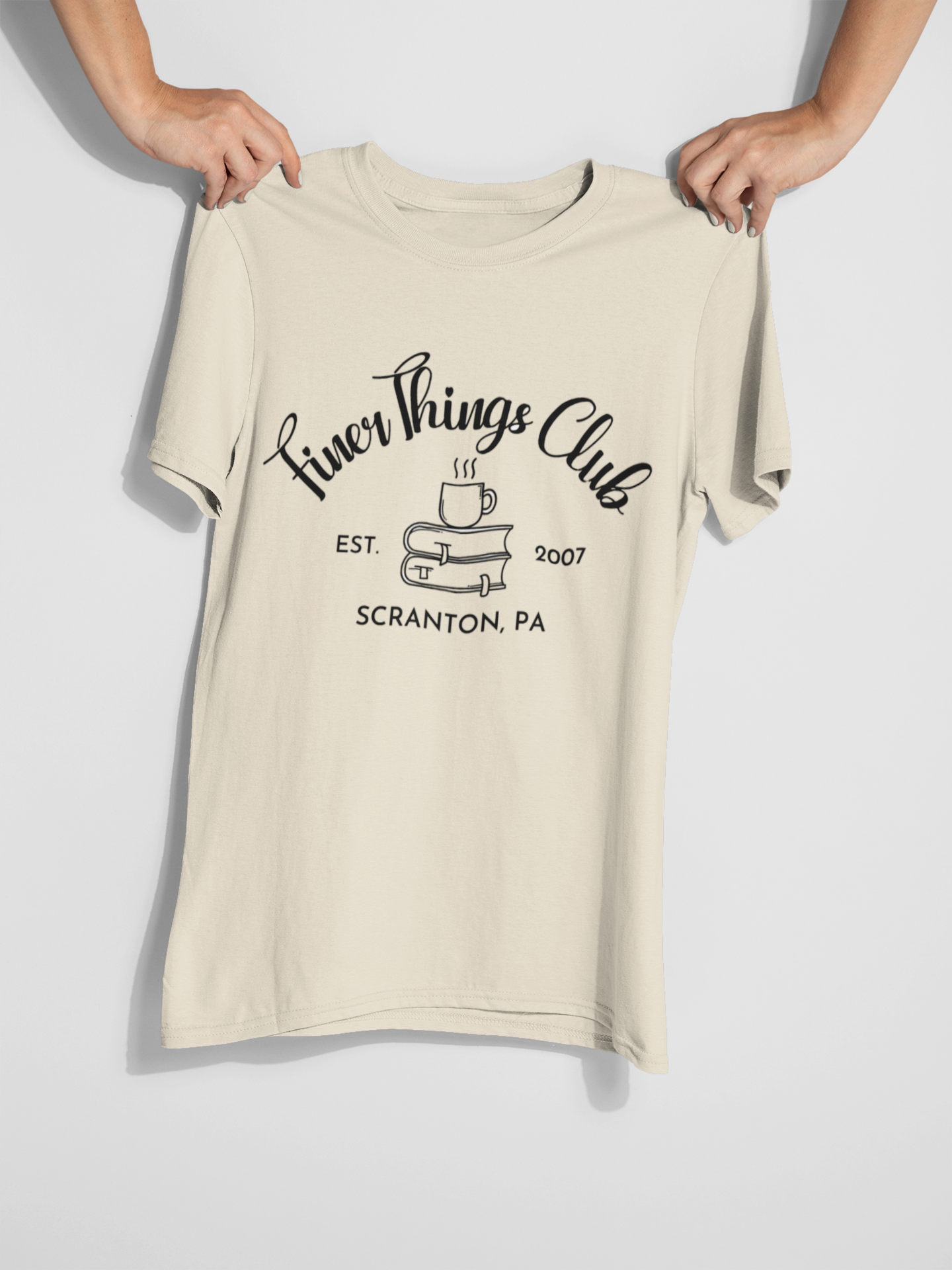 Someone holding a T-Shirt that says "Finer Things Club" with coffee and books on it