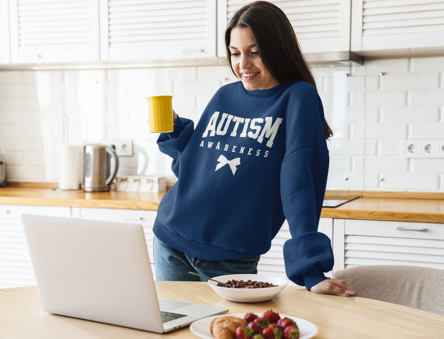 A girl smiling with coffee looking at her laptop wearing a navy sweatshirt that says "Autism Awareness"