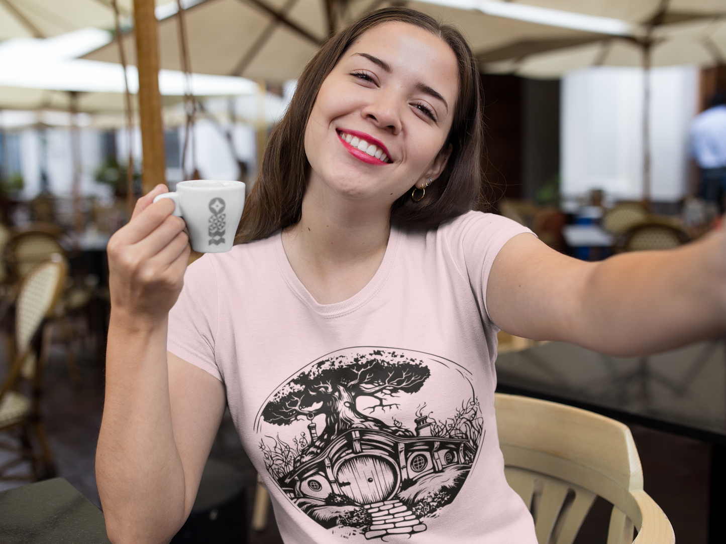 A woman smiling with a cup of coffee wearing a pink T-shirt with a hobbit home on it